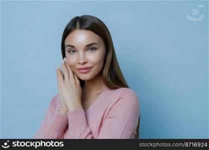 Close up portrait of pleased attractive brunette woman gazes directly at camera, keeps hands near face, listens advice from cosmetologist how to care about skin, models indoor against blue wall