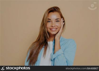 Close up portrait of positive happy woman in white tshirt and blue shirt on beige colored background touches her face tenderly with hand, smiling sincerely on camera. Positive women emotions. Close up portrait of positive happy woman in white tshirt and blue shirt