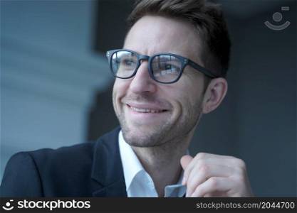 Close up portrait of positive young male entrepreneur in glasses and formal wear looking aside with pleasant smile distracted from remote work on computer in office. Freelance and business concept. Positive young male entrepreneur in glasses and formal wear looking aside with pleasant smile