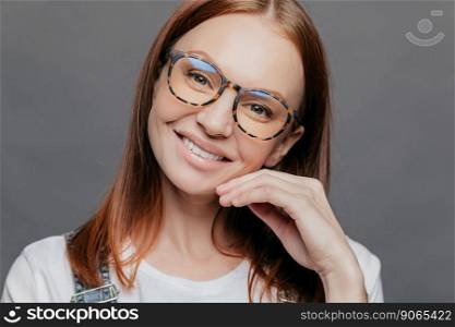 Close up portrait of pretty female model with European appearance, smiles gently, glad to recieve good comments from boss, wears spectacles, keeps hand near face, isolated over grey wall, tilts head