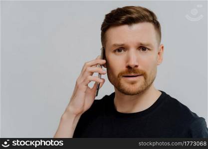 Close up portrait of puzzled young man talking on mobile phone and feeling unpleased after receiving bad news, looking at camera with concerned face expression, standing against grey studio wall. Worried man having unpleasant conversation on mobile phone and feeling unhappy