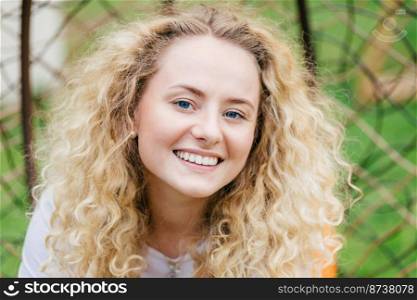 Close up portrait of romantic beautiful fair haired female with appealing smile, rests outdoor, being in good mood, feels inspired by beauty of nature, expresses positive emotions. People, lifestyle
