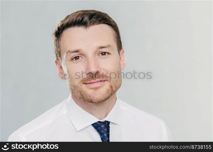 Close up portrait of self assured handsome businessman with stubble, being prosperous business owner, dressed elegantly, stands against white wall. Male boss poses indoor. People and success