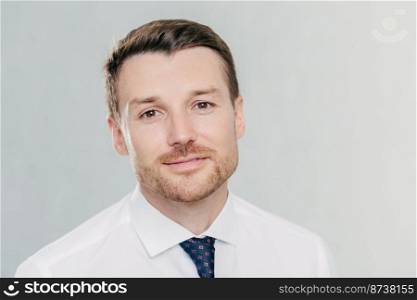 Close up portrait of self assured handsome businessman with stubble, being prosperous business owner, dressed elegantly, stands against white wall. Male boss poses indoor. People and success