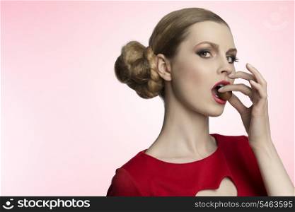 close-up portrait of sensual female with blonde hair-style and cute make-up eating one chocolate praline and wearing red dress