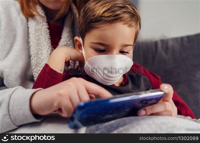 Close up portrait of small boy caucasian male child kid sitting in his mother&rsquo;s lap wearing protective mask antivirus antibacterial pollution disease prevention during pandemic playing on mobile phone