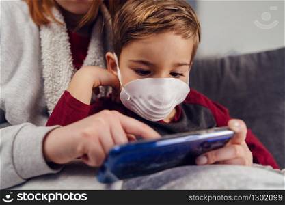 Close up portrait of small boy caucasian male child kid sitting in his mother&rsquo;s lap wearing protective mask antivirus antibacterial pollution disease prevention during pandemic playing on mobile phone