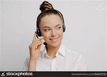 Close-up portrait of smiling female bank worker in white shirt and hair in bun, speaking on black headset, sharing customer information, looking aside, isolated on grey background.
