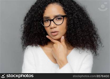 Close up portrait of thoughtful African American woman holds chin, looks mysteriously at camera, has bushy crisp hair, wears optical glasses and white jumper, makes plan, isolated over grey background