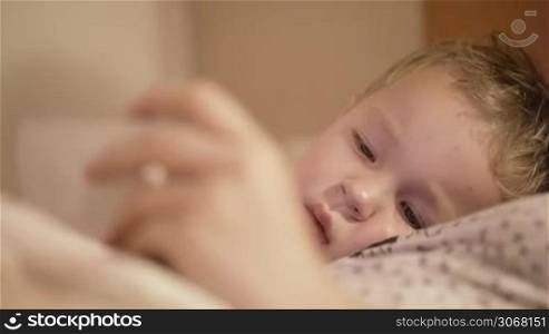 Close up portrait of three year old boy lying on the bed with his mother and watching cartoons on smart phone before he goes to sleep