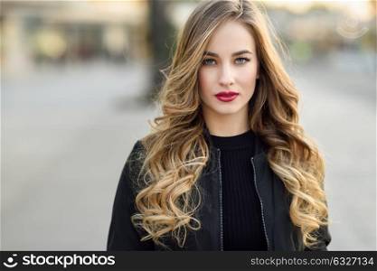 Close-up portrait of young blonde girl with beautiful blue eyes wearing black jacket outdoors. Pretty russian female with long wavy hair hairstyle. Woman in urban background.