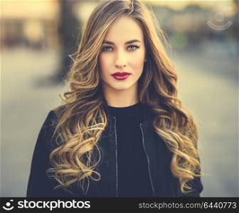 Close-up portrait of young blonde girl with beautiful blue eyes wearing black jacket outdoors. Pretty russian female with long wavy hair hairstyle. Woman in urban background.