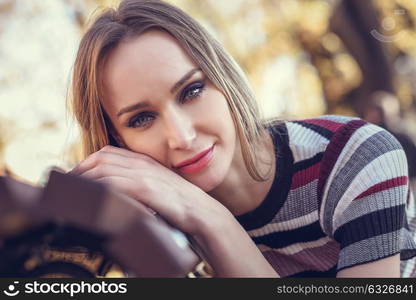 Close-up portrait of young blonde woman sitting on a bench in the street of a park with autumn colors. Beautiful girl with blue eyes in urban background wearing striped dress smiling.