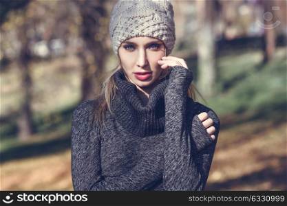 Close-up portrait of young blonde woman standing in a park with autumn colors. Beautiful girl wearing winter gray dress and wool cap. Female with straight hair and blue eyes.