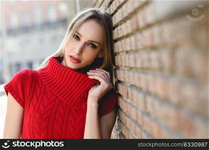 Close-up portrait of young blonde woman standing in the street near a brick wall. Beautiful girl in urban background wearing red dress. Female with straight hair and blue eyes.