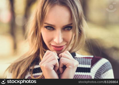 Close-up portrait of young blonde woman with blue eyes standing in the street. Beautiful girl in urban background wearing striped sweater. Female with straight hair.