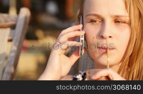 Close up portrait of young caucasian woman talking on her smart phone and drinking coffee coctail with a straw while sitting on the deckchair relaxing on the beach