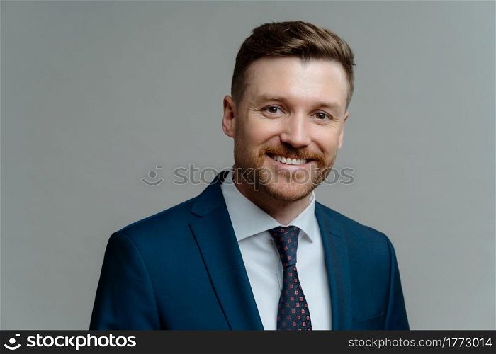 Close up portrait of young cheerful male boss wearing suit smiling at camera, handsome successful businessman or entrepreneur in formal wear posing against grey background. Business people concept. Happy successful entrepreneur in suit smiling at camera while posing against grey background