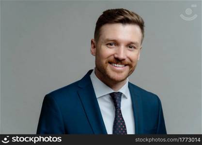Close up portrait of young cheerful male boss wearing suit smiling at camera, handsome successful businessman or entrepreneur in formal wear posing against grey background. Business people concept. Happy successful entrepreneur in suit smiling at camera while posing against grey background