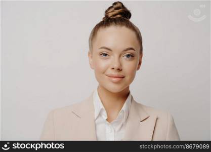 Close up portrait of young pretty brunette business lady with hair bun wearing formal clothes with white shirt standing isolated in front of grey background looking forward with confidence. Confident attractive business lady in beige suit