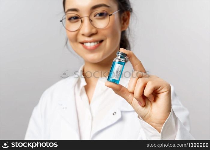 Close-up portrait of young professional asian doctor, researcher or infectionist, giving patient ampule with covid19 vaccine, smiling pleased, starting tests of treatment sick people, grey background.. Close-up portrait of young professional asian doctor, researcher or infectionist, giving patient ampule with covid19 vaccine, smiling pleased, starting tests of treatment sick people, grey background