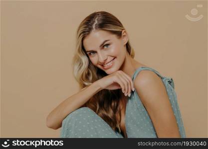 Close up portrait of young sensual tender female wearing summer dress feeling shy while looking at camera, smiling and waiting for romantic date while posing over beige background in studio. Young sensual tender female wearing summer dress feeling shy while looking at camera