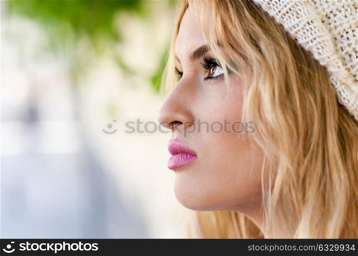 Close-up profile of blonde young woman with curly hair. Girl wearing sweater and wool cap in urban background. Female with beautiful lips painted in pink
