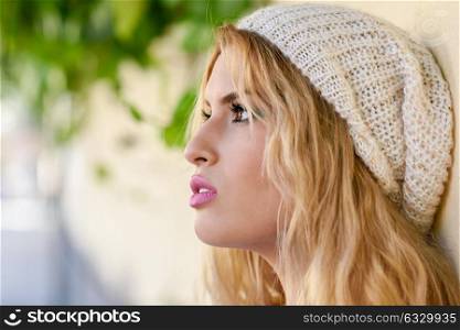 Close-up profile of blonde young woman with curly hair. Girl wearing sweater and wool cap in urban background. Female with beautiful lips painted in pink