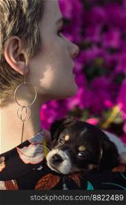 Close up puppy on shoulder of young woman in garden portrait picture. Closeup rear view photography with flowers on background. High quality photo for ads, travel blog, magazine, article. Close up puppy on shoulder of young woman in garden portrait picture