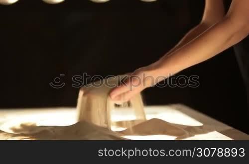 Close-up sand spilling out of female hands against black background in dimmed lights. Artist drawing on the sand, creating sand painting. Hands of a female master creating artwork making sand animation.