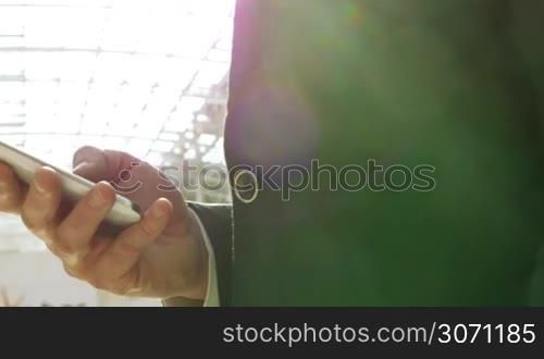 Close-up shot of a businessman using smart phone to type sms or playing game in bright sunlight coming through the transparent roof of modern office building or trade center