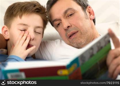 Close-up shot of a father reading a book to the son who is already sleepy and rubbing his eyes. Bedtime reading with father
