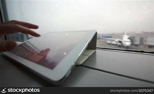 Close-up shot of a female hand typing on tablet computer by the window at airport on background of boarding plane