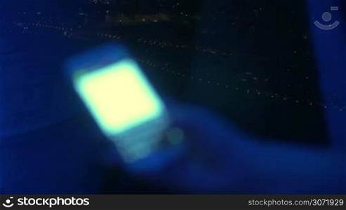 Close-up shot of a female hand typing sms or email on smart phone on the background of distant city lights at night. At first focus on the city view then on the cell phone