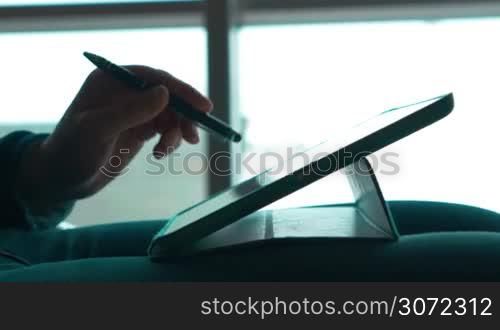 Close-up shot of a female hands working with tablet computer using pen to touch screen. She holding tablet PC on the lap agaisnt the window with bright sunlight