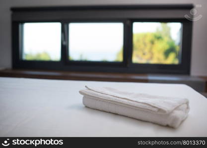 Close-up shot of a fresh white towel on the bed in hotel room, window with nature scene in background