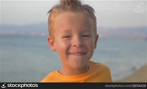 Close-up shot of a little boy in orange t-shirt with mohawk on the head looking to the camera and smiling. Happy careless child against sea background