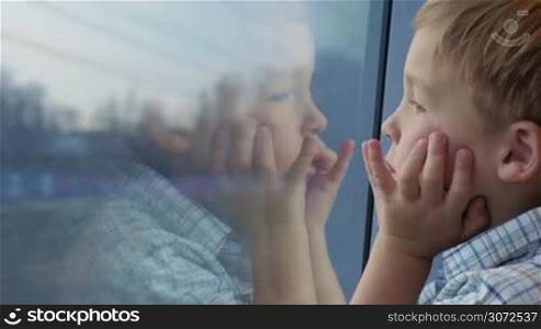 Close-up shot of a little boy traveling by train and looking out the window. His hands on the face and reflection in the glass