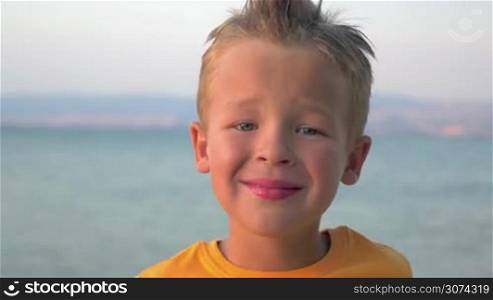 Close-up shot of a little cute boy with mohawk hair-do smiling. Portrait against blue sea and sky background