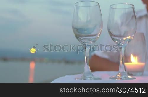 Close-up shot of a man pouring white wine into two glasses standing on the table with candle in outdoor restaurant on the beach