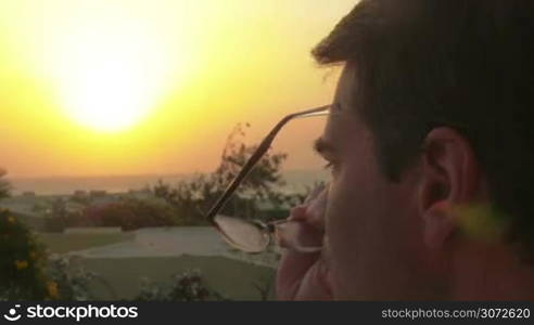 Close-up shot of a man putting on glasses and looking at beautiful sunset over the sea on resort