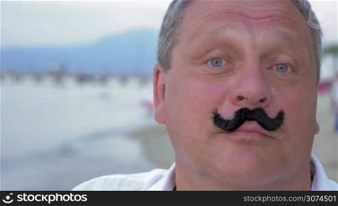 Close-up shot of a mature man touching up fake mustache with funny face expression. Shot on the beach
