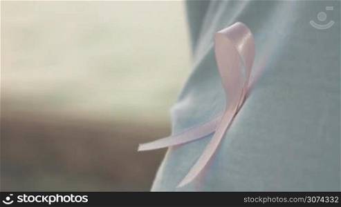 Close-up shot of a person wearing pink breast cancer awareness ribbon on white t-shirt, defocused sea in background