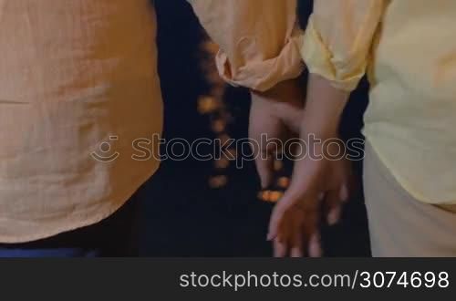 Close-up shot of a senior man and woman joining and holding hands by the dark sea water sparkling with light reflection at night