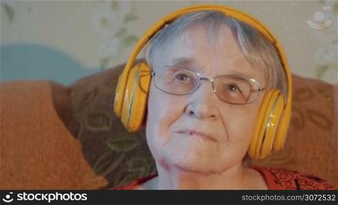 Close-up shot of a senior woman in glasses enjoying music in wireless headphones at home. Leisure of aged people