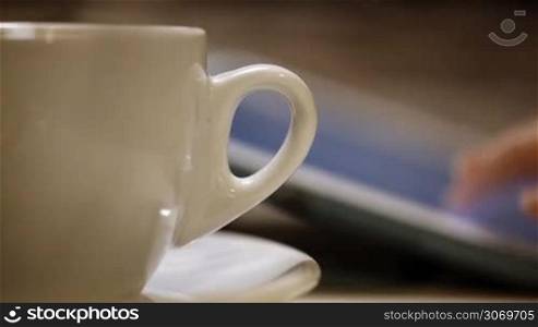 Close-up shot of a woman having a cup of coffee during using tablet PC. Only hand, cup and pad in shot. Focus on the cup of coffee