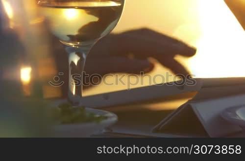 Close-up shot of a woman in outdoor cafe on the shore at sunset. She typing message on tablet computer and having drink. Sea in golden colors of sunset in background