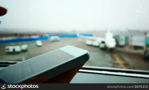 Close-up shot of a woman typing message on smart phone by the window at the airport, plane in background