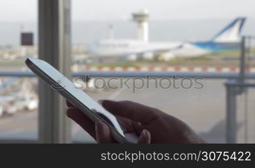 Close-up shot of a woman typing sms on smart phone by the window of airport terminal with view to the moving airplane