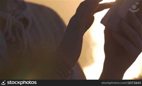Close-up shot of a woman working with touch pad against bright golden sunset reflecting in water